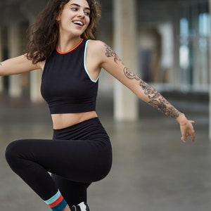 May include: A woman wearing a black athletic crop top with a blue and orange trim, black leggings with colorful stripes at the ankle, and black and white sneakers. She is smiling and has a tattoo on her arm.
