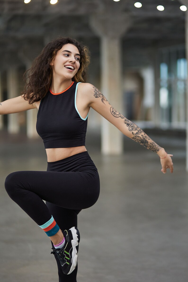 May include: A woman wearing a black athletic crop top with a blue and orange trim, black leggings with colorful stripes at the ankle, and black and white sneakers. She is smiling and has a tattoo on her arm.