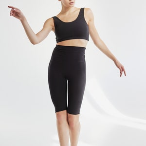K&ouml;nnte beinhalten: Eine Frau tr&auml;gt ein schwarzes &auml;rmelloses Crop-Top und schwarze, hochgeschlossene Radlerhosen. Sie steht auf einem wei&szlig;en Hintergrund und schaut nach rechts.