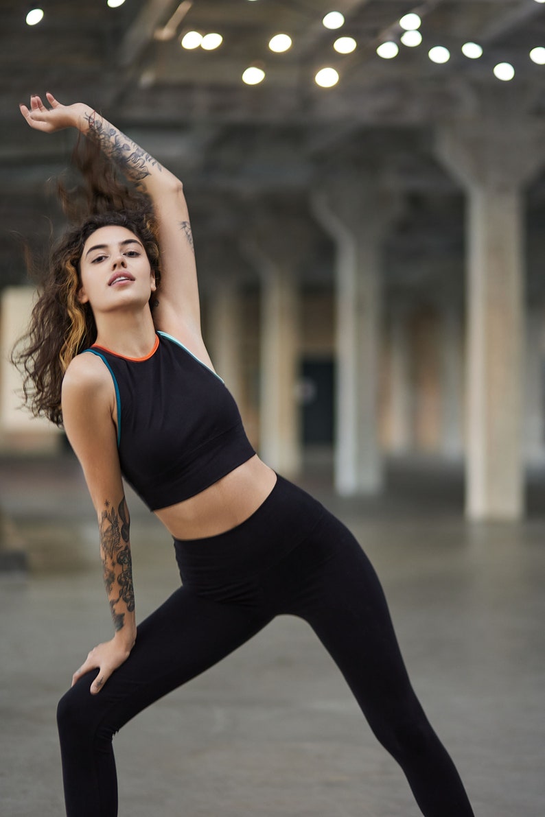May include: A woman wearing a black sports bra with orange and blue trim and black leggings poses in a warehouse setting.