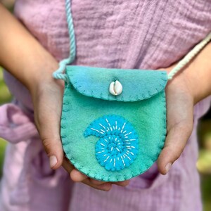 May include: A small, round, teal felt purse with a seashell closure and a turquoise spiral design. The purse has a matching teal strap. The purse is held by a person wearing a pink shirt.