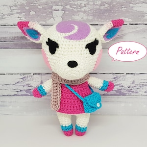 May include: A crocheted animal plush toy resembling a white deer with pink cheeks, purple and blue ears, and a pink dress. The toy is wearing a brown scarf and a blue bag. The text "Pattern" is written in a speech bubble.