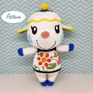 Puede incluir: Muñeca de oveja hecha a ganchillo, predominantemente blanca con ojos negros, nariz rosa y detalles azules y amarillos. Tiene un vestido con bordados florales. La palabra "Pattern" está en una burbuja.