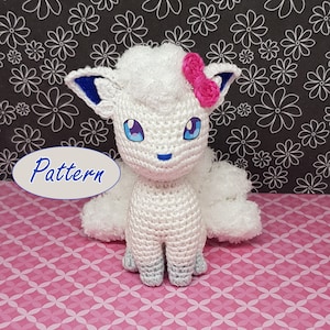 Könnte beinhalten: Weißes gehäkeltes Amigurumi-Spielzeug eines Fuchses mit blauen Ohren, blauen Augen und einer rosa Schleife. Das Spielzeug sitzt auf einer rosa-weißen gemusterten Oberfläche. Das Wort "Pattern" ist in einem weißen Oval auf blauem Hintergrund geschrieben.