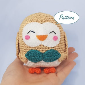 May include: A crocheted owl plush toy with a beige body, white face, orange beak, and green leaves around its neck. The owl has a happy expression with pink cheeks and closed eyes. The word "Pattern" is written in a speech bubble above the owl.