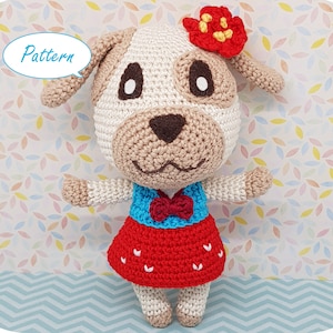 Könnte beinhalten: Ein gehäkeltes Hundespielzeug mit einem weißen und braunen Gesicht, einem roten und weißen Kleid und einer roten Blume auf dem Kopf. Das Spielzeug steht auf einem gemusterten Hintergrund.