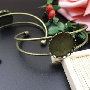 May include: Two antique bronze-toned metal cuff bracelets with circular bezel settings. The bracelets have a curved design with decorative details. The image also includes decorative elements like flowers and greenery.
