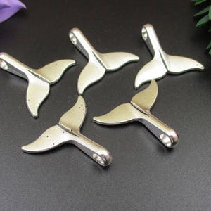 Puede incluir: Seis abalorios de cola de ballena de metal plateado. Los abalorios miden aproximadamente 5 cm de largo y tienen un bucle en la parte superior para sujetarlos a un collar o pulsera.
