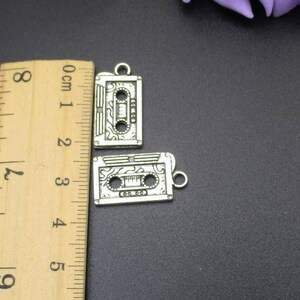 May include: Two silver-toned cassette tape charms with a detailed design. The charms are approximately 2 cm in length and are shown next to a ruler.