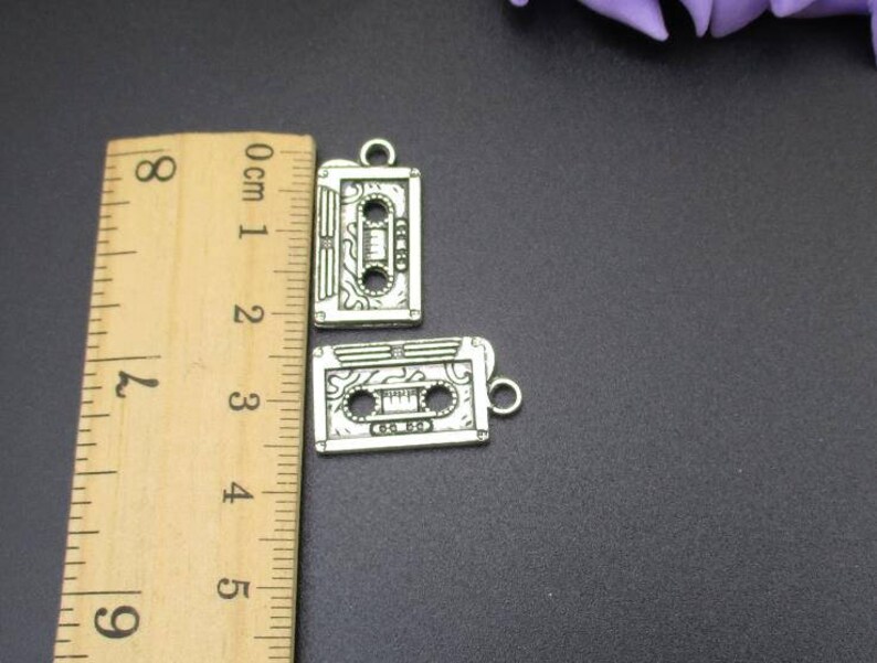 May include: Two silver-toned cassette tape charms with a detailed design. The charms are approximately 2 cm in length and are shown next to a ruler.