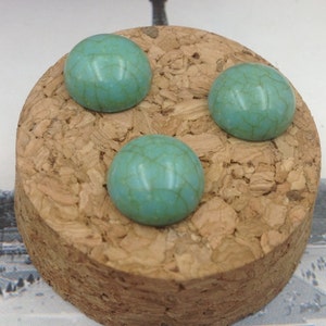 May include: Three turquoise blue cabochon stones with a marbled pattern are displayed on a cork surface.