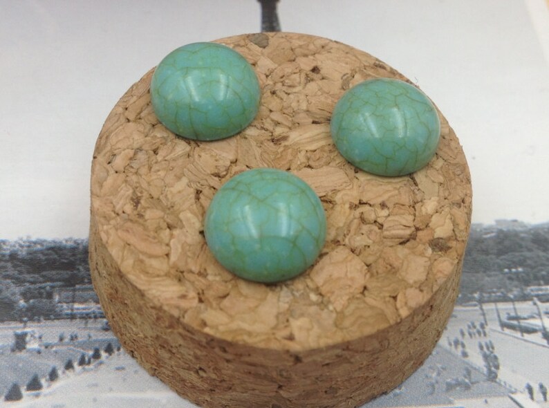 May include: Three turquoise blue cabochon stones with a marbled pattern are displayed on a cork surface.