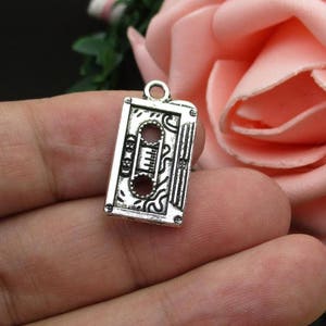 May include: Silver charm in the shape of a vintage cassette tape. The charm has an intricate design and is perfect for adding a retro touch to jewelry.