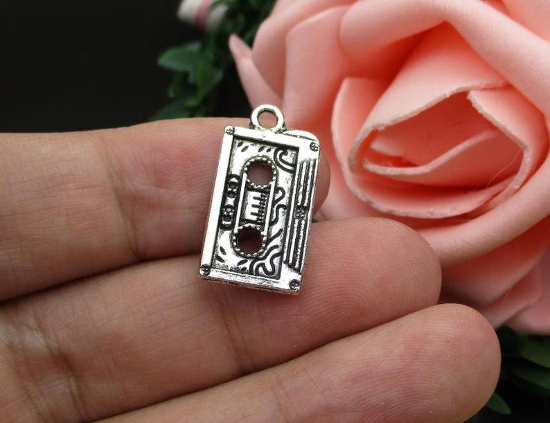 May include: Silver charm in the shape of a vintage cassette tape. The charm has an intricate design and is perfect for adding a retro touch to jewelry.