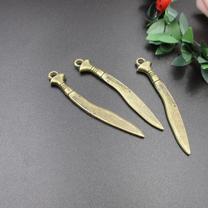 Puede incluir: Tres abalorios de espada de color dorado antiguo con un diseño detallado. Los abalorios están sobre una superficie gris oscura.