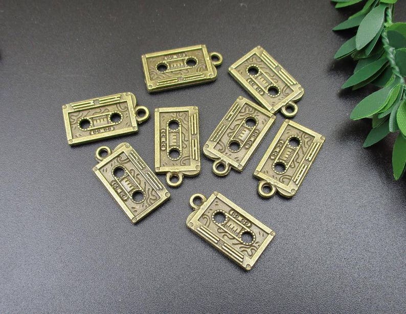 Puede incluir: Una colecci&oacute;n de ocho abalorios de metal de color dorado antiguo con forma de cintas de casete. Cada abalorio tiene un dise&ntilde;o detallado y un bucle para sujetarlo a un collar, pulsera o llavero.
