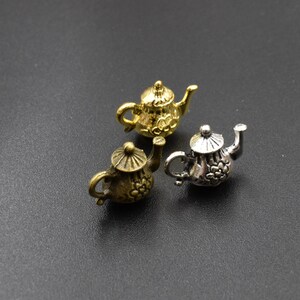 Può includere: Tre teiere in miniatura, una dorata, una argentata e una di bronzo, con intricati disegni floreali. Le teiere sono su uno sfondo nero.