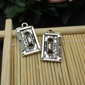 May include: Two silver-toned metal charms shaped like vintage cassette tapes. Each charm has a detailed design with a decorative pattern and a raised outline of the cassette tape.