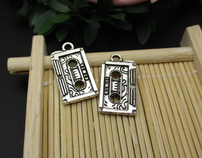 May include: Two silver-toned metal charms shaped like vintage cassette tapes. Each charm has a detailed design with a decorative pattern and a raised outline of the cassette tape.
