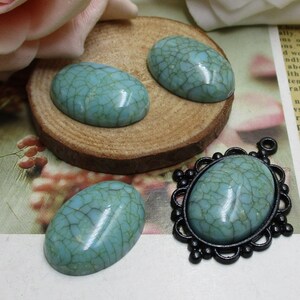 May include: Three oval-shaped turquoise cabochons with a crackled finish. The cabochons are on a wooden surface. One cabochon is in a black metal setting with a decorative border.