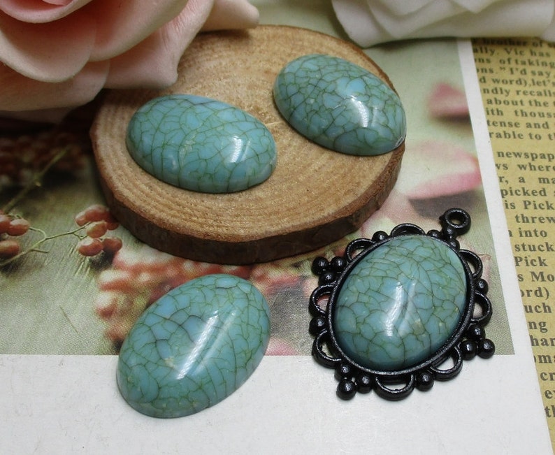 May include: Three oval-shaped turquoise cabochons with a crackled finish. The cabochons are on a wooden surface. One cabochon is in a black metal setting with a decorative border.