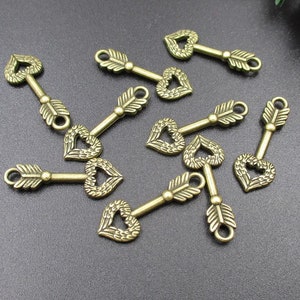 Puede incluir: Un grupo de diez dijes de llave de color dorado antiguo con mirillas en forma de corazón y tallos de llave en forma de flecha. Los dijes están esparcidos sobre una superficie gris oscuro.