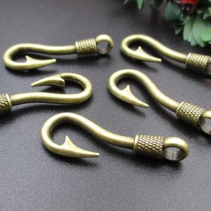 Op de afbeelding: Vijf antieke goudkleurige metalen vishaakbedels met een getextureerd ontwerp. De haken zijn allemaal in dezelfde richting gericht en zijn in een halve cirkel gerangschikt.