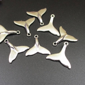 May include: Silver-toned metal charms in the shape of mermaid tails, each with a small loop at the top for attaching to jewelry or other crafts. The charms are scattered on a dark surface.