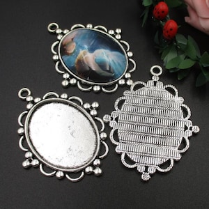 May include: Three silver-toned metal pendant settings for jewellery making. Two of the settings are oval shaped with a decorative border. The third setting is round with a more intricate design.