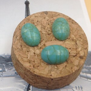 May include: Three turquoise oval cabochons with a marbled pattern, displayed on a cork surface.