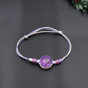 Puede incluir: Una pulsera hecha a mano con un diseño floral redondo púrpura en resina transparente. La pulsera tiene un cordón blanco y lavanda con dos pequeñas cuentas púrpuras a cada lado de la pieza central floral. Fondo oscuro.