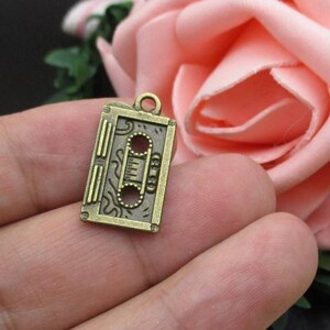 Puede incluir: Un amuleto de metal de color bronce con forma de cinta de casete. El amuleto tiene un dise&ntilde;o detallado con un patr&oacute;n ondulado y una forma rectangular.