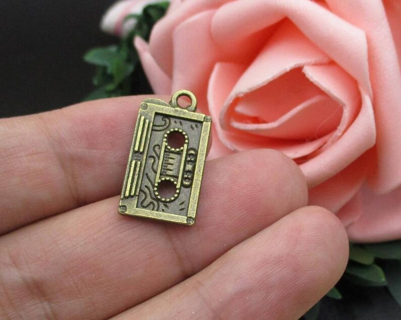 Puede incluir: Un amuleto de metal de color bronce con forma de cinta de casete. El amuleto tiene un dise&ntilde;o detallado con un patr&oacute;n ondulado y una forma rectangular.