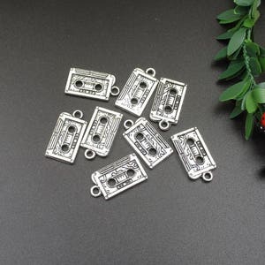 May include: Eight silver-toned metal charms shaped like cassette tapes. Each charm has a detailed design of a cassette tape with a black and white print.