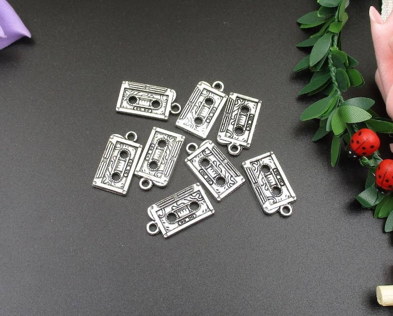 May include: Eight silver-toned metal charms shaped like cassette tapes. Each charm has a detailed design of a cassette tape with a black and white print.