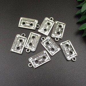 May include: Eight silver-toned metal charms shaped like cassette tapes. Each charm has a detailed design of a cassette tape with a black and white print. The charms have a loop at the top for attaching to a necklace or bracelet.