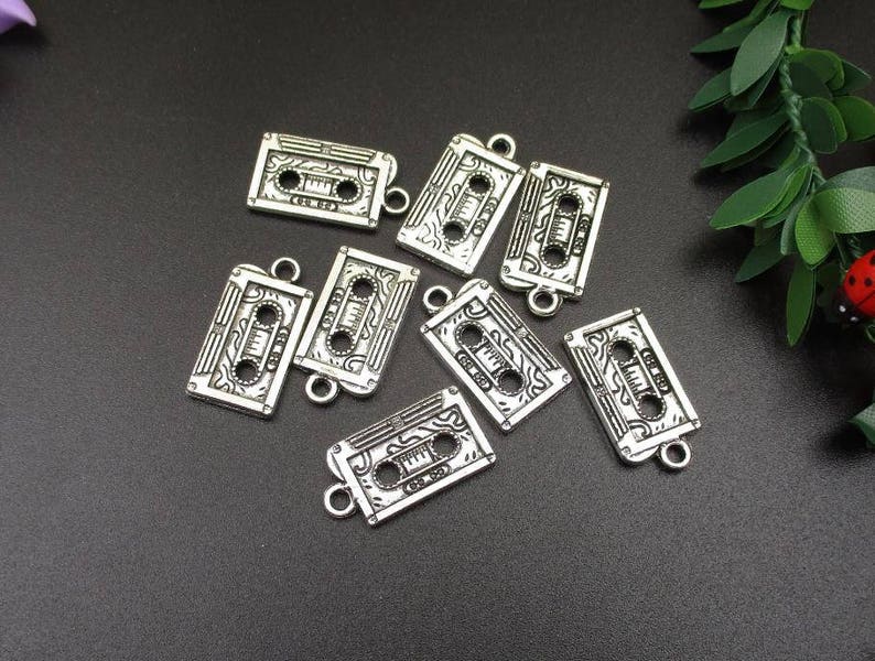 May include: Eight silver-toned metal charms shaped like cassette tapes. Each charm has a detailed design of a cassette tape with a black and white print. The charms have a loop at the top for attaching to a necklace or bracelet.