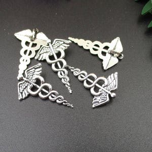 May include: Five silver-toned metal charms in the shape of a caduceus, the symbol of medicine. Each charm features a snake wrapped around a staff with wings.