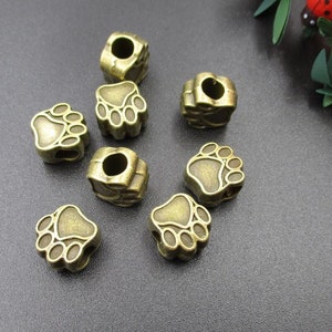May include: A set of eight antique gold-toned metal charms shaped like paw prints. The charms have a hole in the top for attaching to a bracelet or necklace.