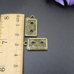 Puede incluir: Dos encantos de metal de color dorado antiguo con forma de cintas de cassette. Los encantos tienen un dise&ntilde;o detallado y miden aproximadamente 2 cm de largo.