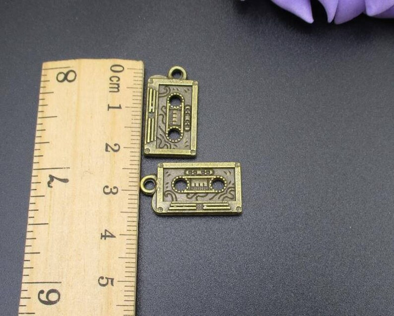 Puede incluir: Dos encantos de metal de color dorado antiguo con forma de cintas de cassette. Los encantos tienen un dise&ntilde;o detallado y miden aproximadamente 2 cm de largo.