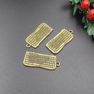 May include: Three antique gold-toned metal charms shaped like computer keyboards. Each charm has a different layout of keys with letters and numbers. The charms have a loop at the top for attaching to a necklace or bracelet.