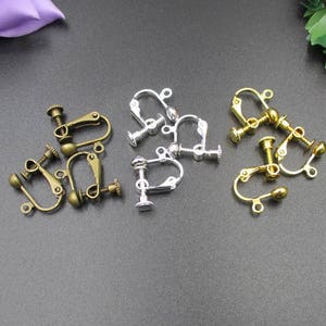 Op de afbeelding: Een assortiment clip-on oorbelonderdelen in antiek messing, zilver en goudtinten. De onderdelen hebben een schroefsluiting en een kleine lus voor het bevestigen van oorbellen. De afbeelding toont verschillende benodigdheden voor het maken van sieraden.