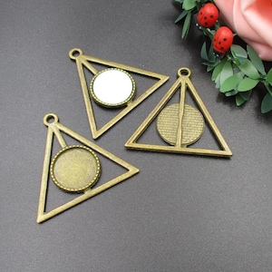 Pode incluir: Três pingentes em forma de triângulo de cor dourada antiga com um cabochão redondo de vidro no centro. Os pingentes têm um design decorativo em torno do vidro.