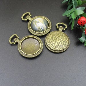 Pode incluir: Três pingentes de cor bronze antigo com um design decorativo. Os pingentes têm uma forma redonda com uma borda elevada e um pequeno laço na parte superior. Um pingente tem um design floral, outro tem um espaço em branco para uma imagem e o terceiro tem uma imagem de um cavalo branco.