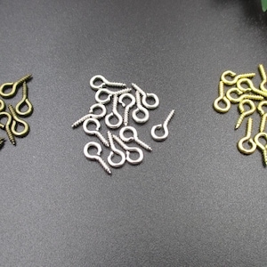 May include: A close-up of three piles of screw-in eye hooks. The piles are arranged in a row from left to right. The left pile is bronze, the middle pile is silver, and the right pile is gold.