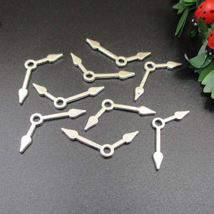 Może przedstawiać: Kolekcja srebrnych wskazówek zegara, każda ze strzałkową końcówką i centralnym otworem, rozrzuconych na ciemnej powierzchni. Wskazówki te są prawdopodobnie przeznaczone do projektu zegara DIY lub naprawy zegarków.