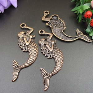 Op de afbeelding: Drie antieke bronzenen zeemeermincharms met ingewikkelde details. Elke charme heeft een zeemeermin met golvend haar en een schubbige staart. De charmes zijn perfect voor het maken van sieraden of knutselen.