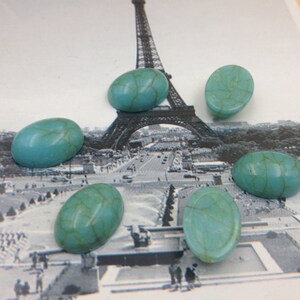 May include: Six oval-shaped turquoise cabochons with a marbled pattern. The cabochons are arranged in a semi-circle on a white background with a black and white image of the Eiffel Tower in the background.
