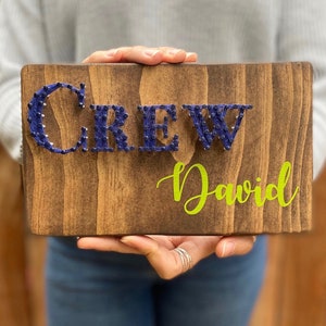 May include: A wooden sign with the word "CREW" spelled out in blue yarn wrapped around nails. The name "David" is written in green below the word "CREW".